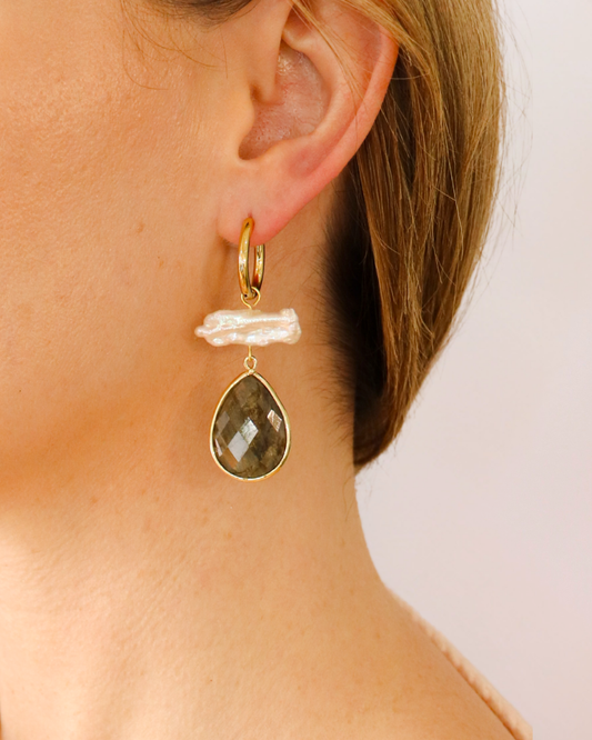 A close-up of a model's ear wearing a gold huggie hoop earring with a textured white baroque pearl and a faceted teardrop-shaped smoky gray gemstone.