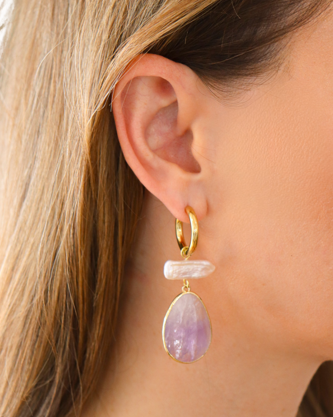 A close-up of a model's ear wearing a gold hoop earring with a dangling light pink stone and a larger teardrop-shaped lavender amethyst drop.